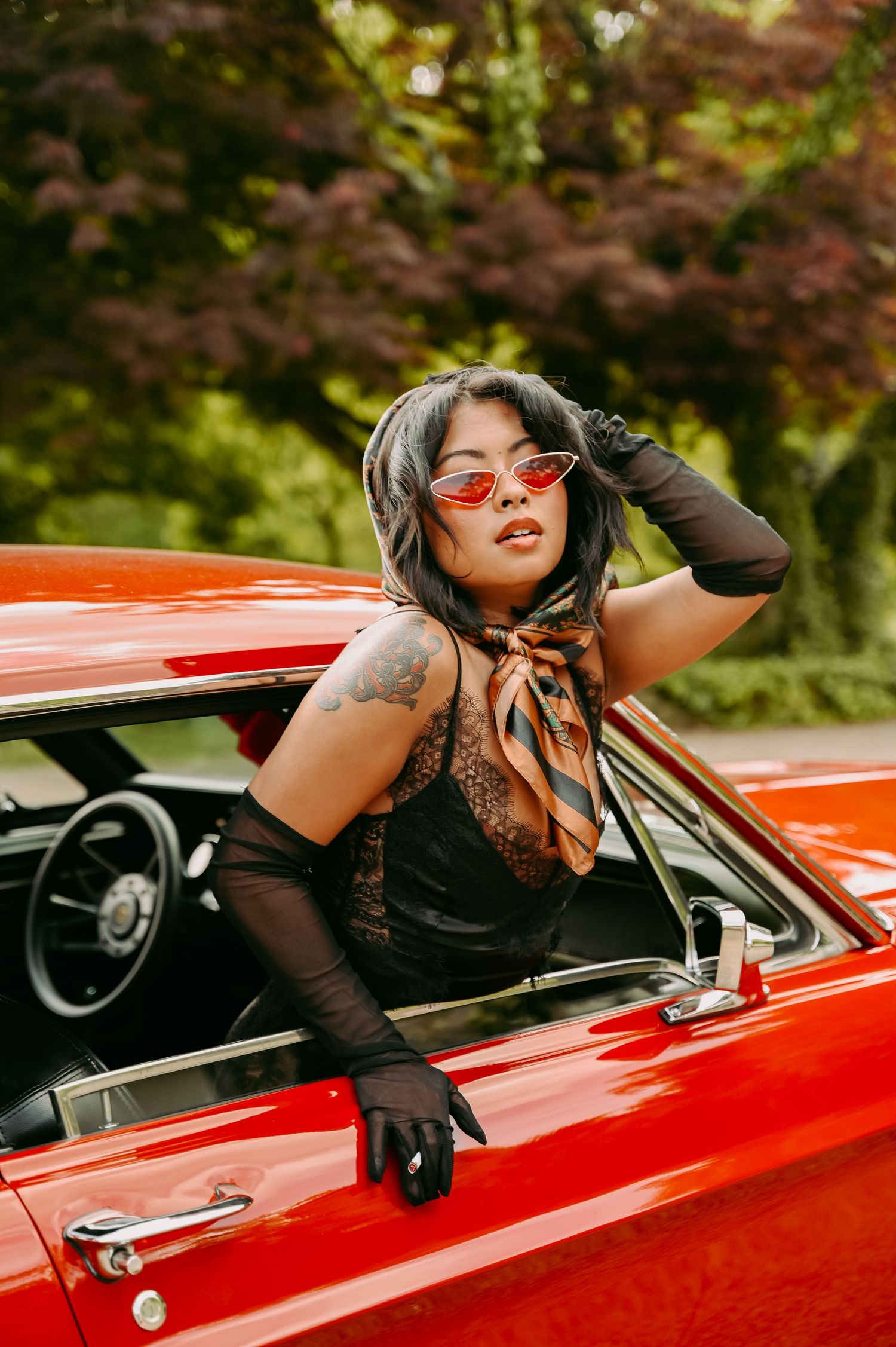 Woman in red sunglasses and black gloves posing in a classic red convertible car surrounded by lush green trees.