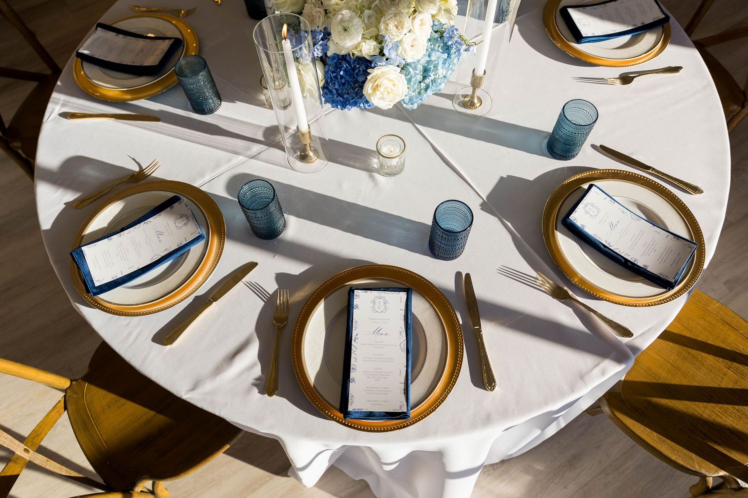 Elegant wedding table setting with gold chargers, blue glassware, white linens, and floral centerpiece.