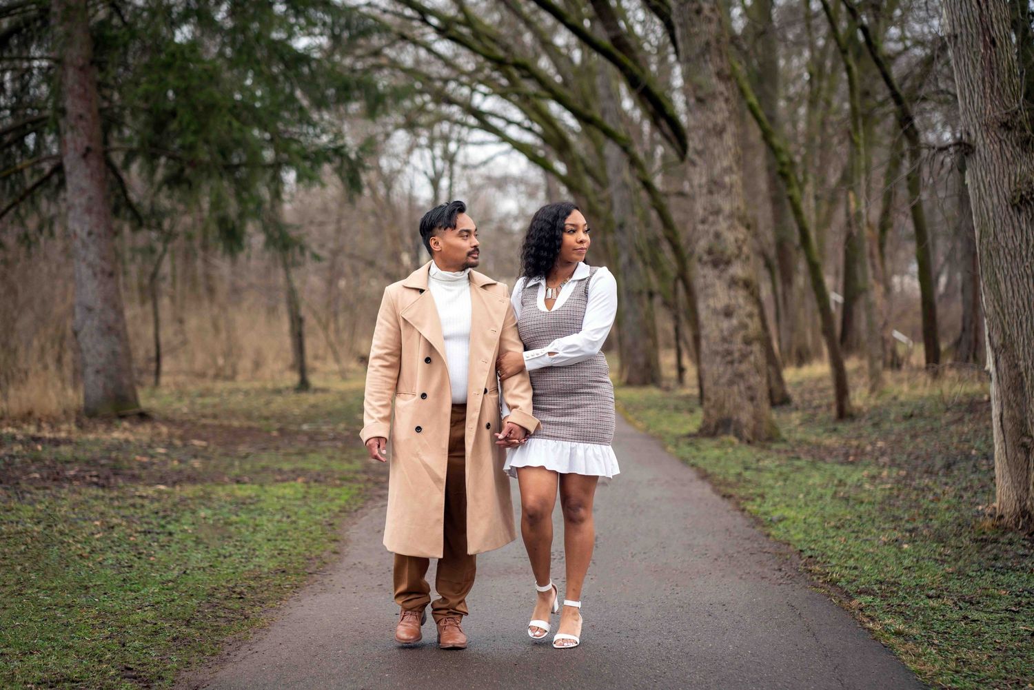 Couple walking hand-in-hand on a tree-lined path in a park, dressed in elegant fall outfits.