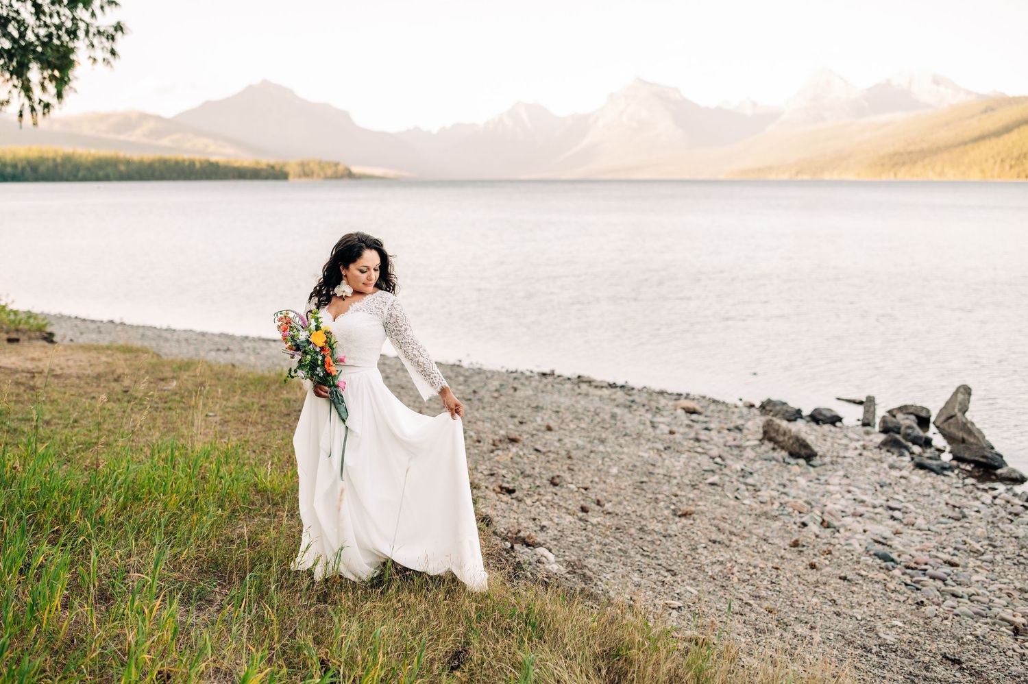 Bride in white lace gown holding colorful bouquet by mountain lake shore on her wedding day.