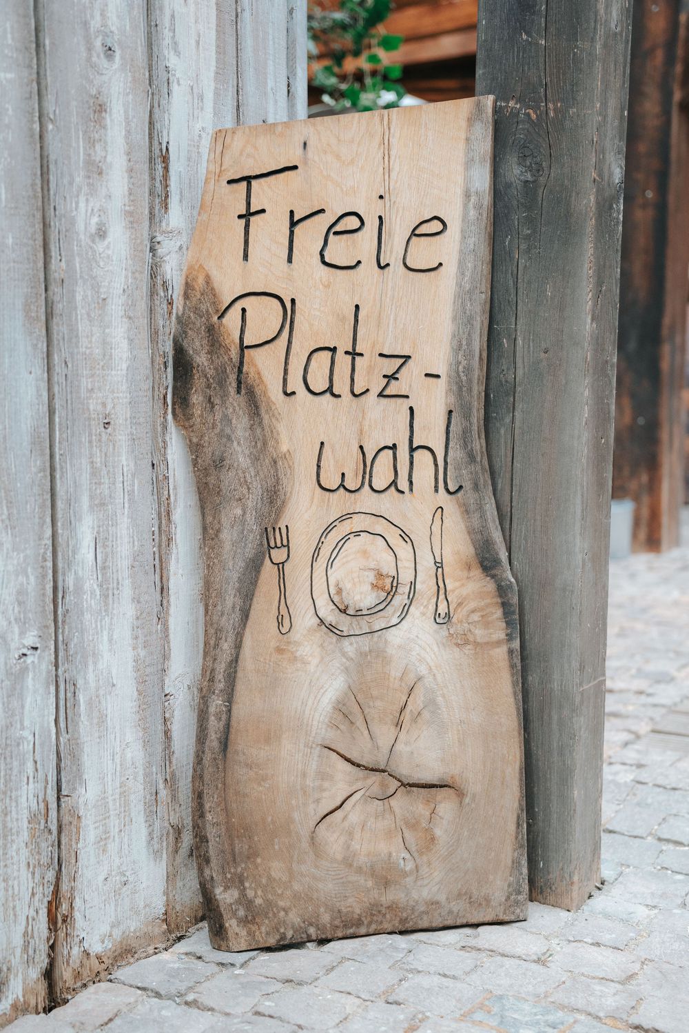 Rustic wooden sign with burned text reading Freie Platzwahl, meaning free seating, with fork, plate, and knife icons.