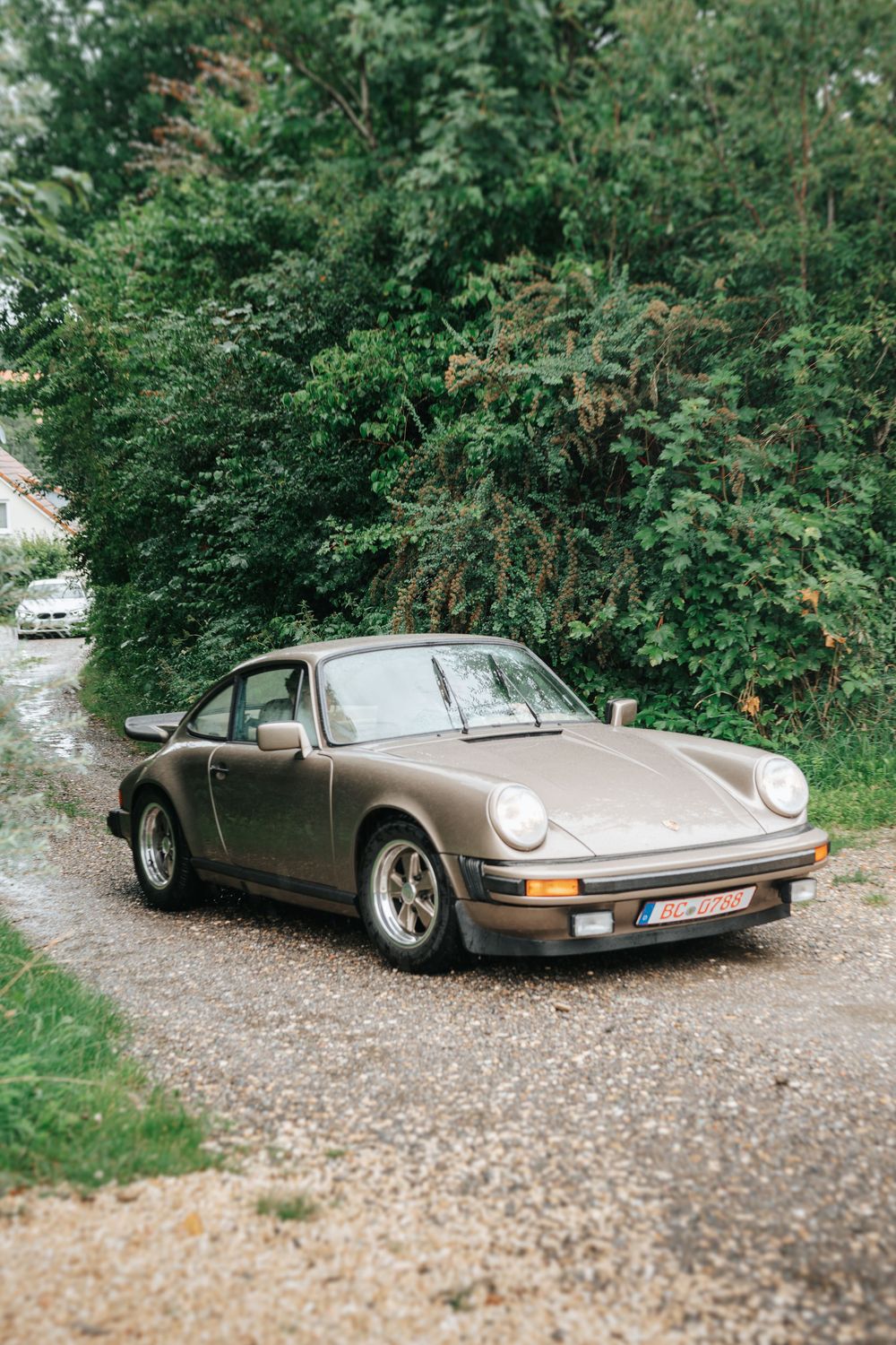 Vintage bronze Porsche 911 parked on a gravel path surrounded by lush green trees.