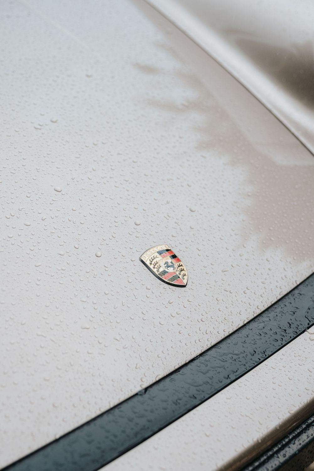 Close-up of a Porsche emblem on a wet white car hood with water droplets and dark racing stripes.