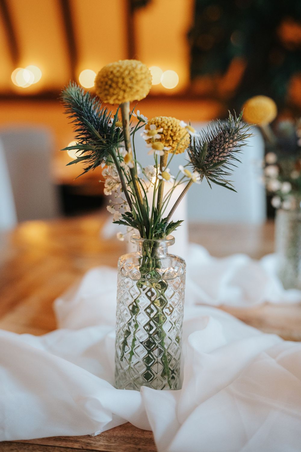Crystal vase with yellow craspedia and blue thistle flowers on white fabric table runner at elegant event.