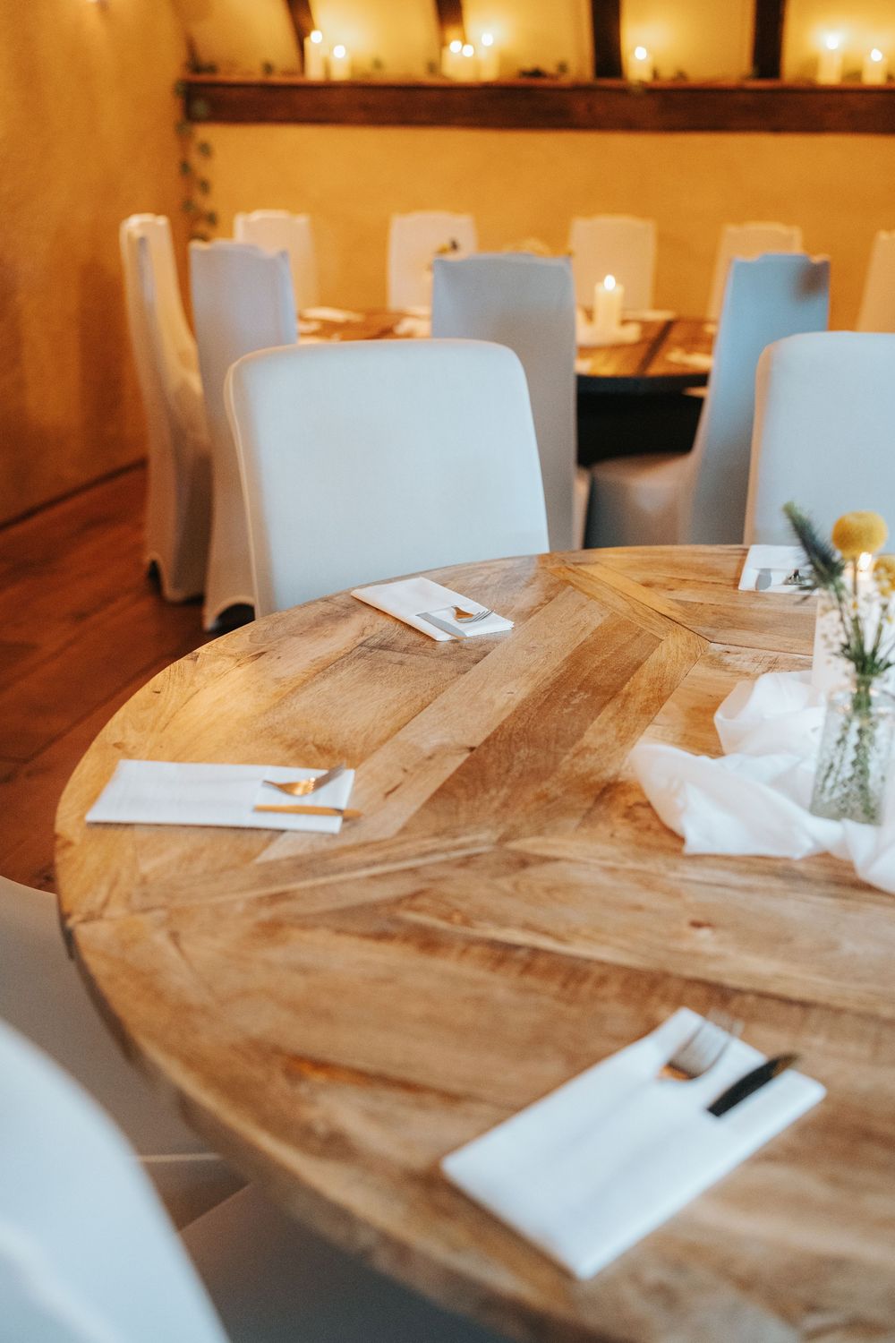Elegant round wooden dining table set with white napkins and cutlery in a warmly lit restaurant.