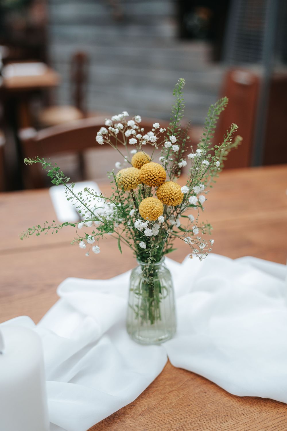 Small glass vase with yellow craspedia and white gypsophila wildflowers on a wooden table with white linen cloth.