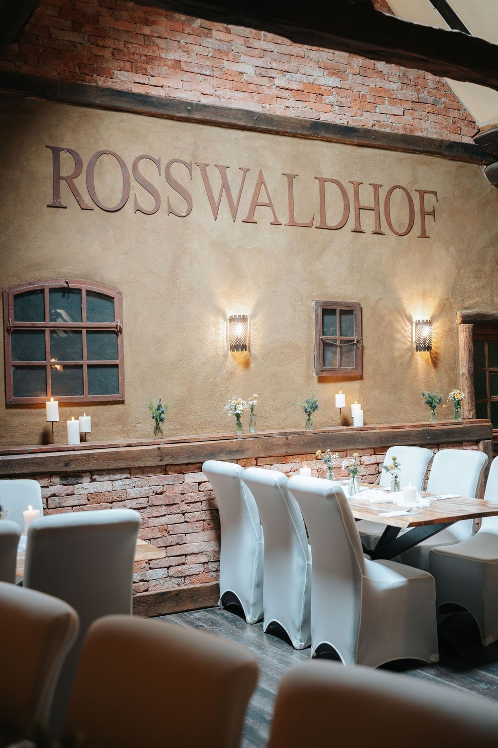 Rosswaldhof restaurant interior with white chairs, candlelit tables, brick walls, and warm wall sconces.