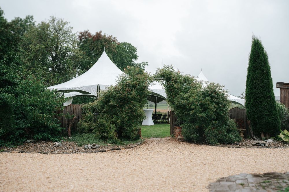 White stretch tents visible through lush green garden archway on a gravel path at an outdoor venue.