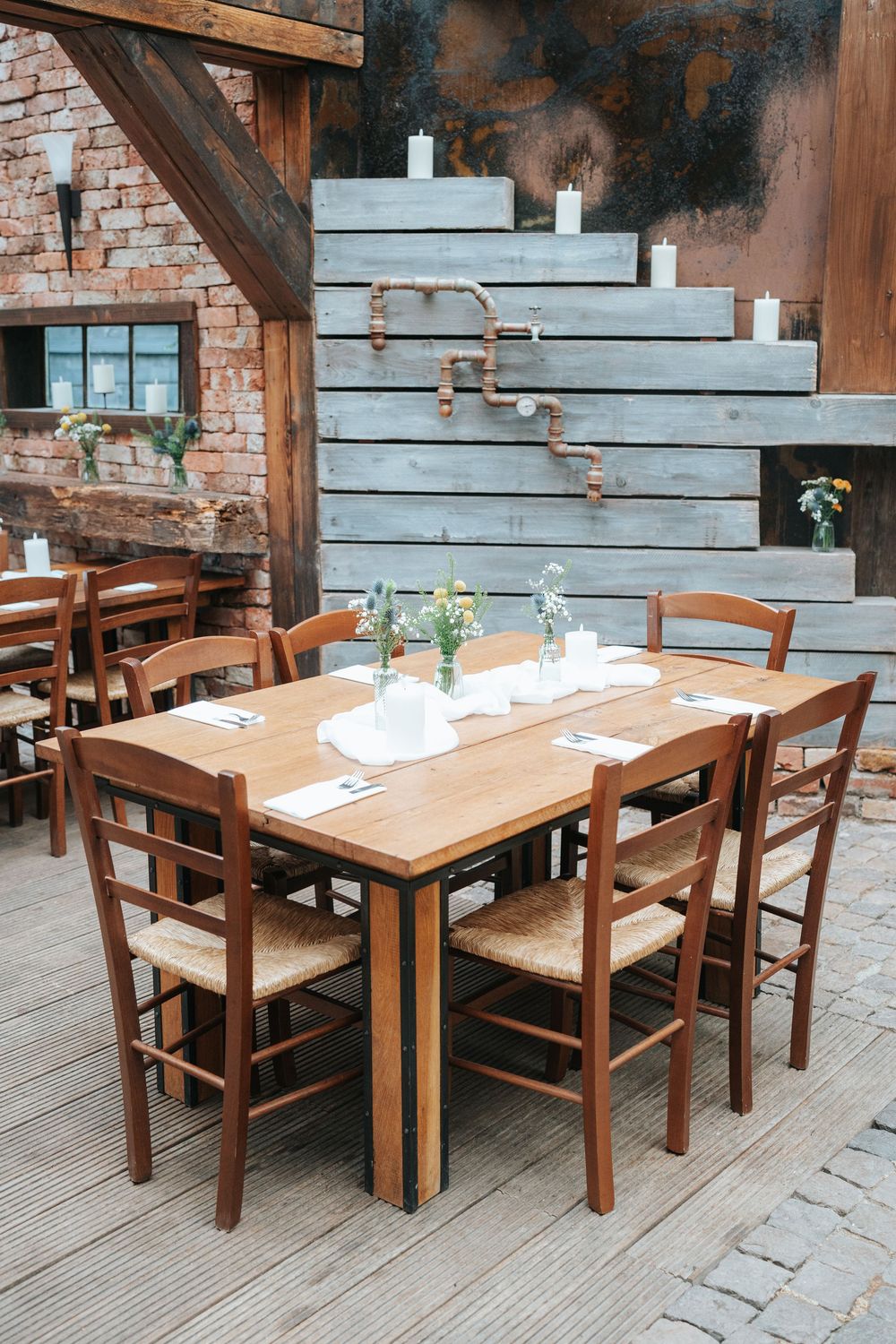 Rustic wooden dining table set with chairs, white napkins, and flower vases in an industrial brick restaurant.