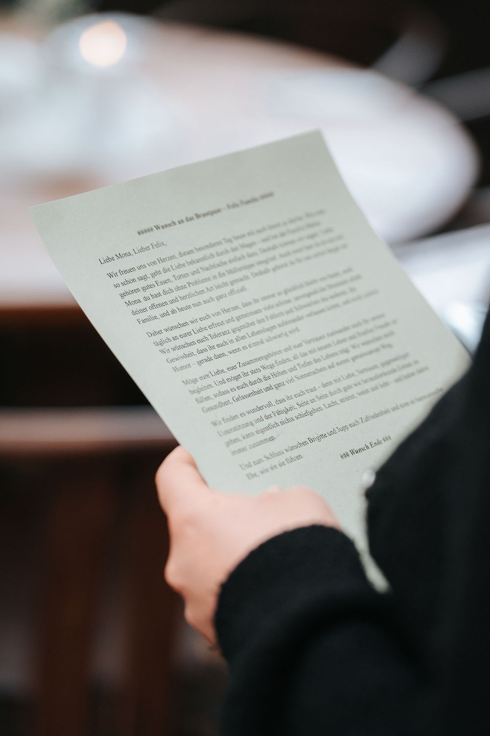 Person in black jacket holding a letter or menu card, reading printed text at a restaurant or event.