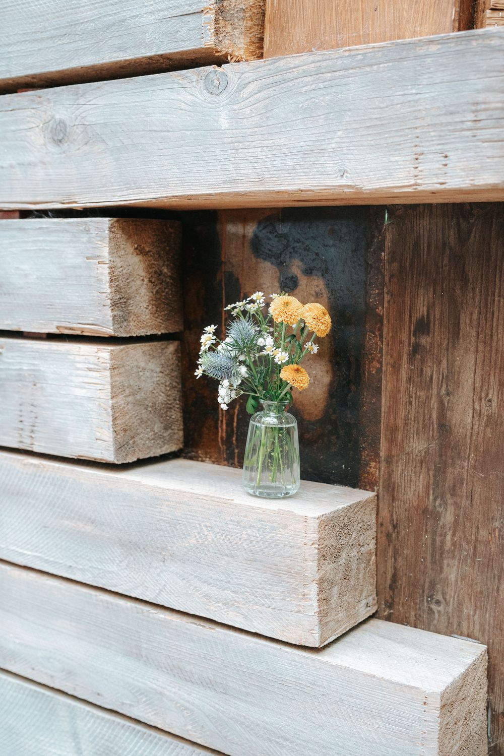 A small glass vase with wildflowers sits on a rustic wooden shelf built into a weathered plank wall.