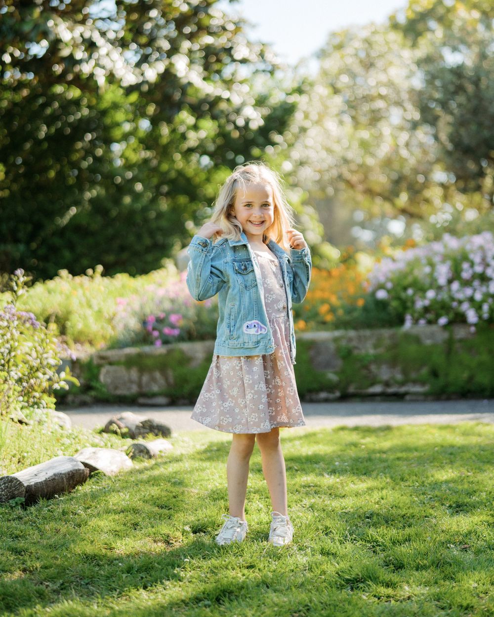 A cheerful young blonde girl in a denim jacket and floral dress poses in a colorful garden.