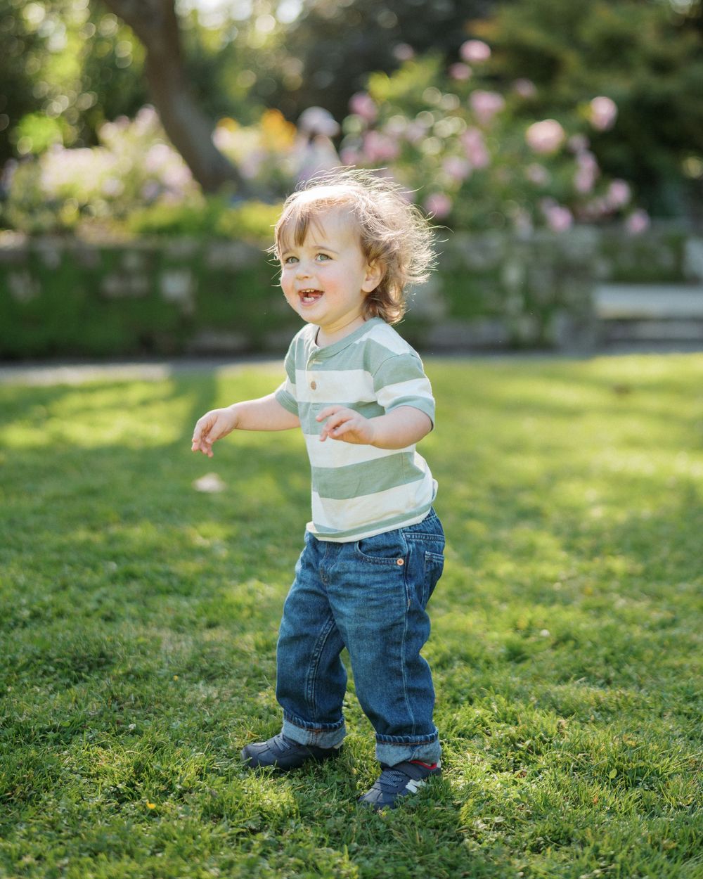 Happy toddler boy in striped shirt walking on green grass in a sunny garden setting.
