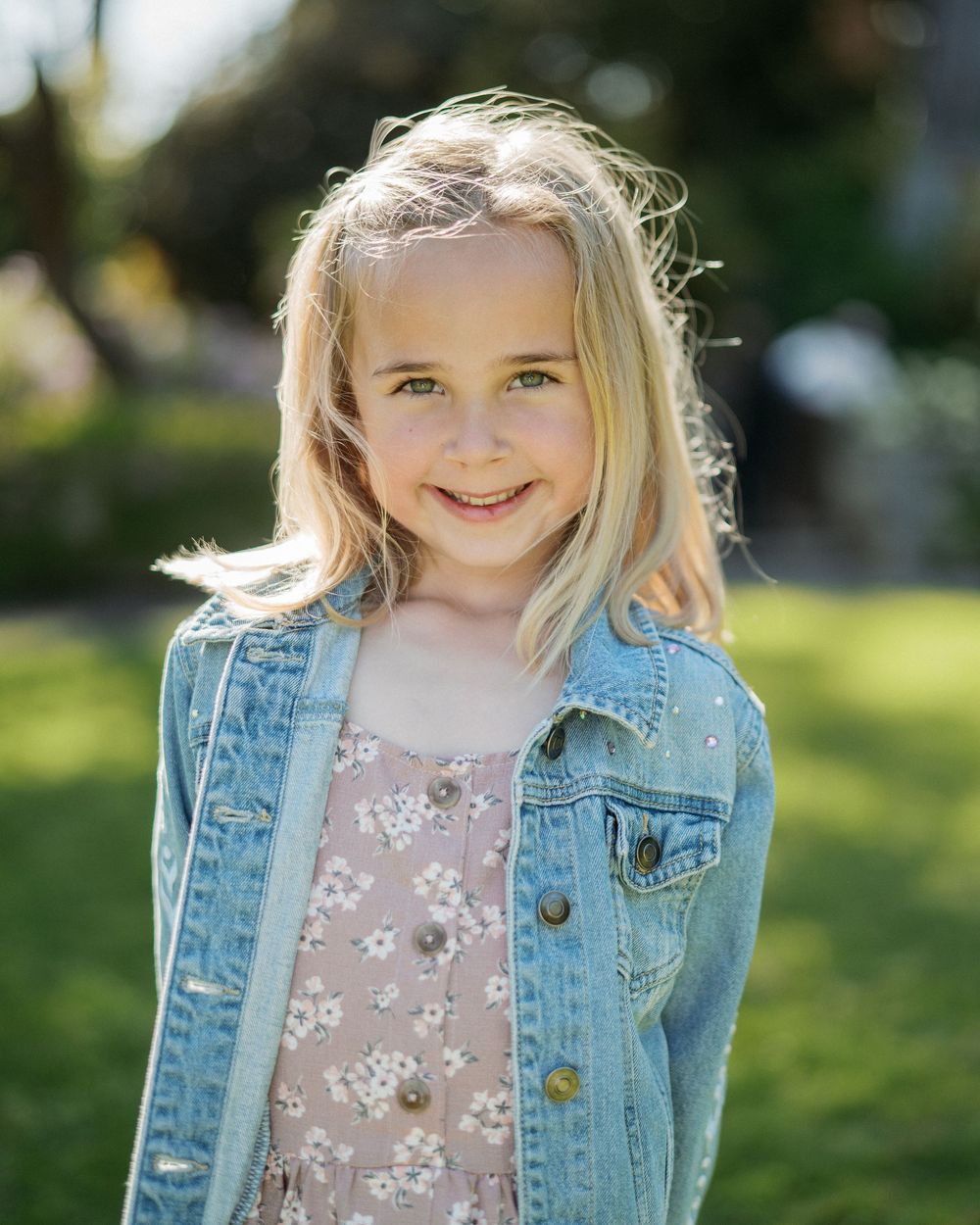 A smiling blonde girl in a denim jacket and floral dress stands in a sunny green outdoor setting.