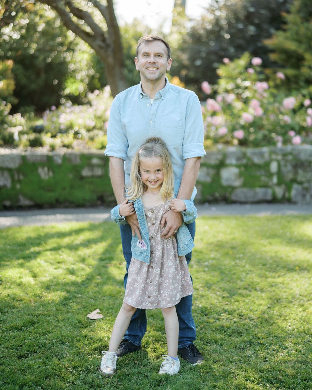 Smiling father standing with young blonde daughter in a garden with pink roses blooming.