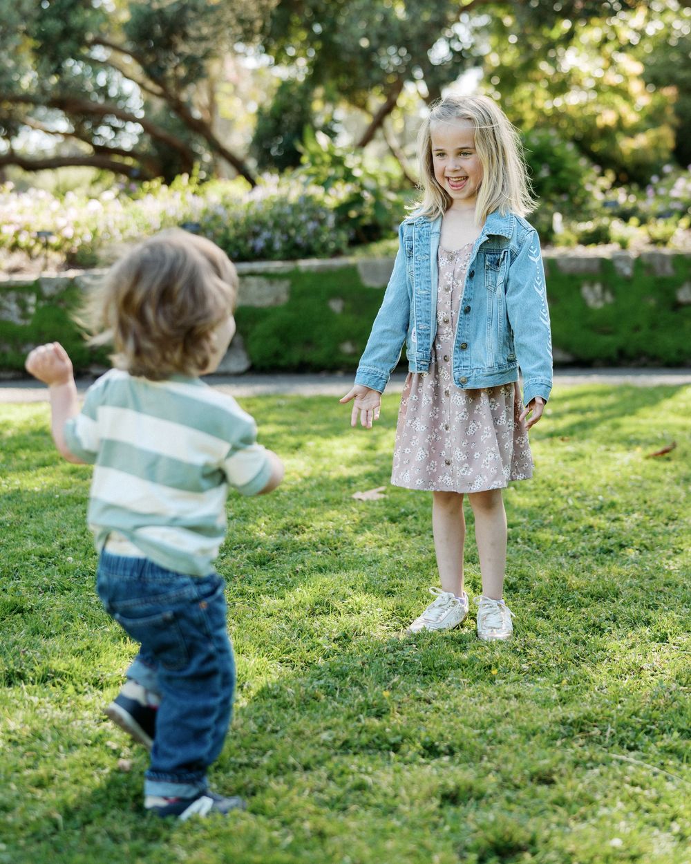 Two young children play together on green grass in a sunny garden, smiling and reaching toward each other.