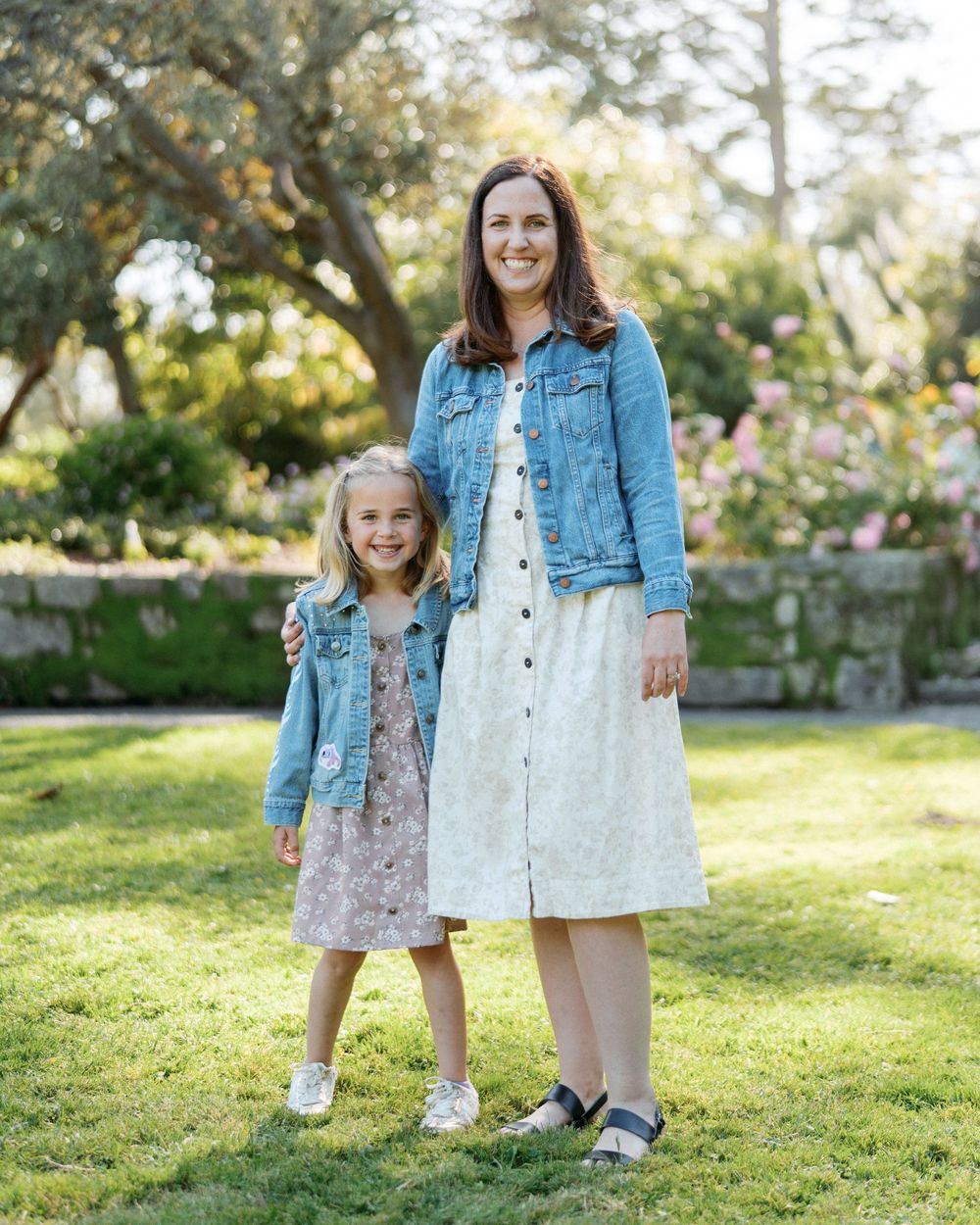 A mother and young daughter in matching denim jackets smile together on a sunny lawn.