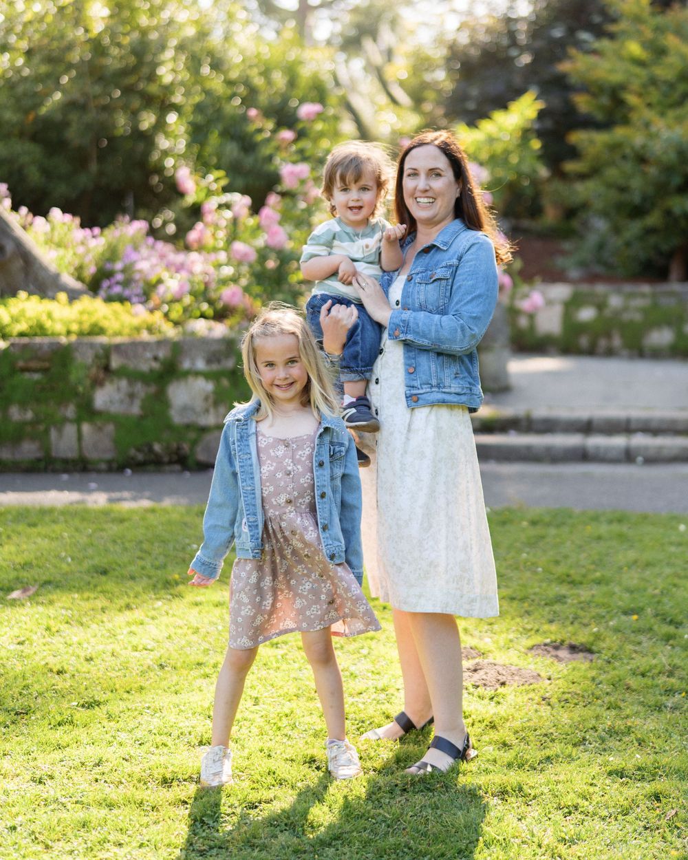 Mother holding toddler son while standing with blonde daughter in a sunny garden setting.