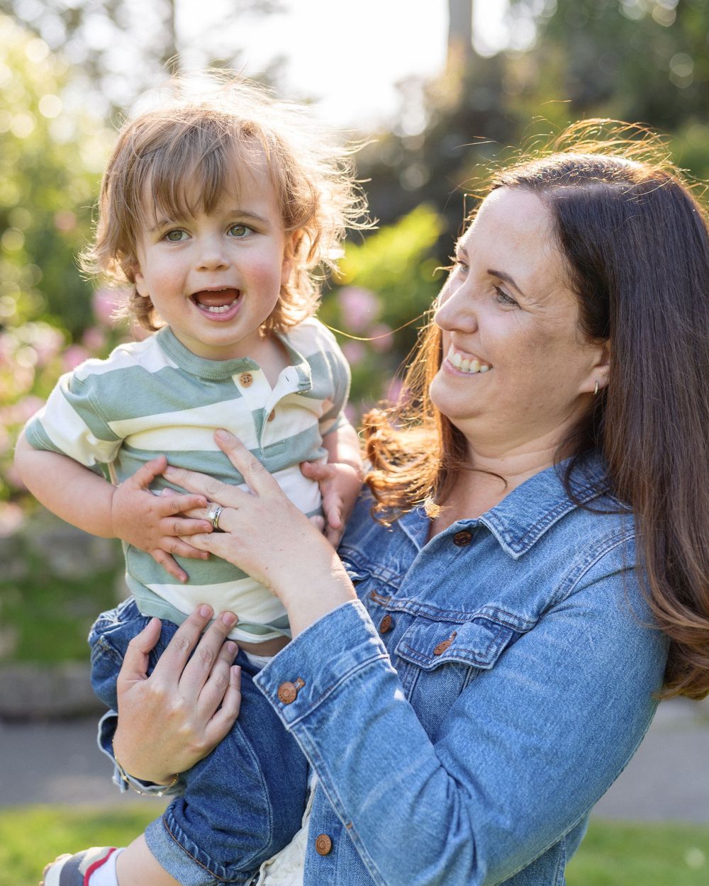 A smiling woman in a denim shirt holds a happy toddler boy outdoors in a sunlit garden with pink flowers.