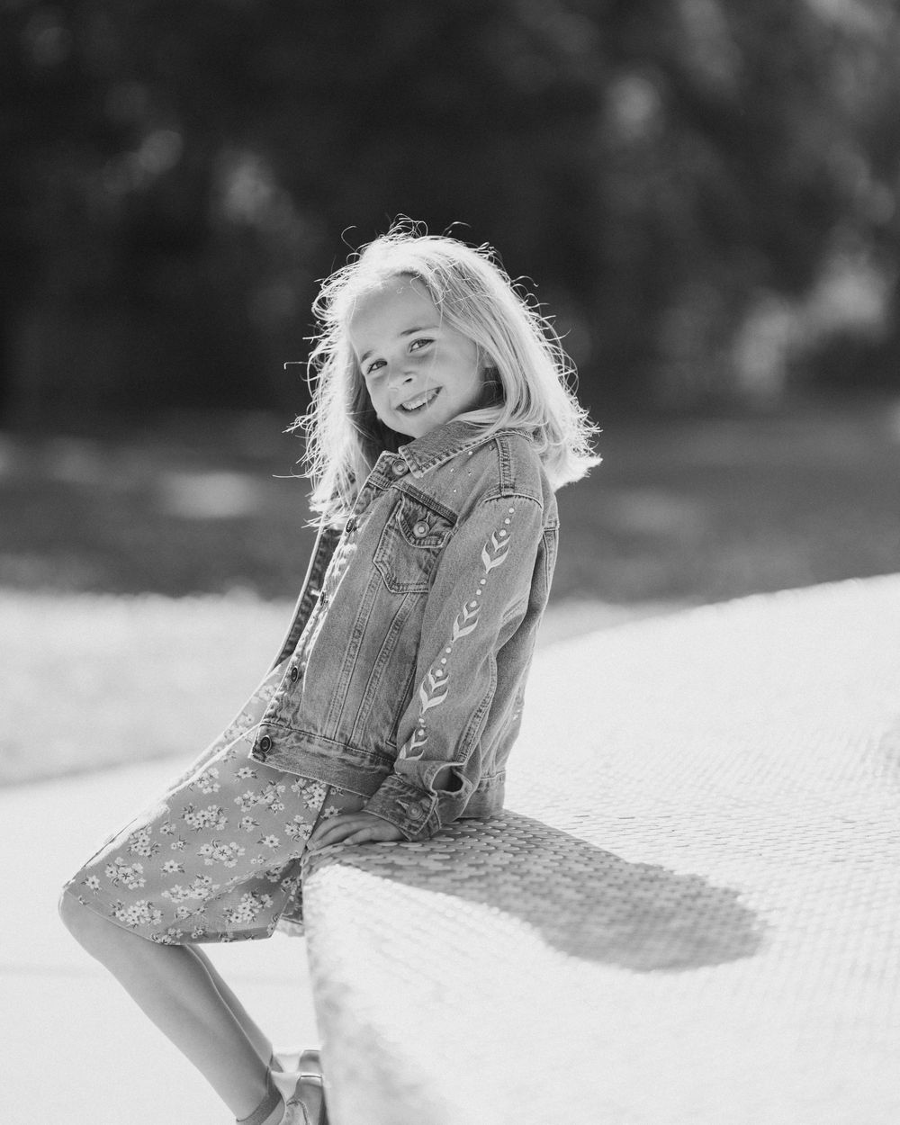 Black and white portrait of a smiling blonde girl in a denim jacket and floral dress posing outdoors.