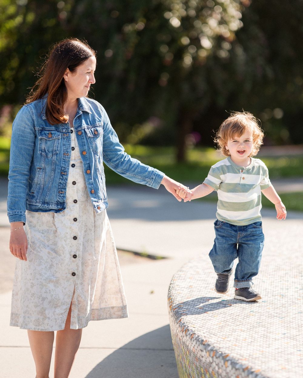 A mother in a denim jacket and floral skirt holds hands with her toddler son outdoors.