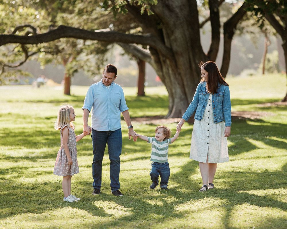 Family of four holding hands and walking on green grass in a sunny park with large trees.