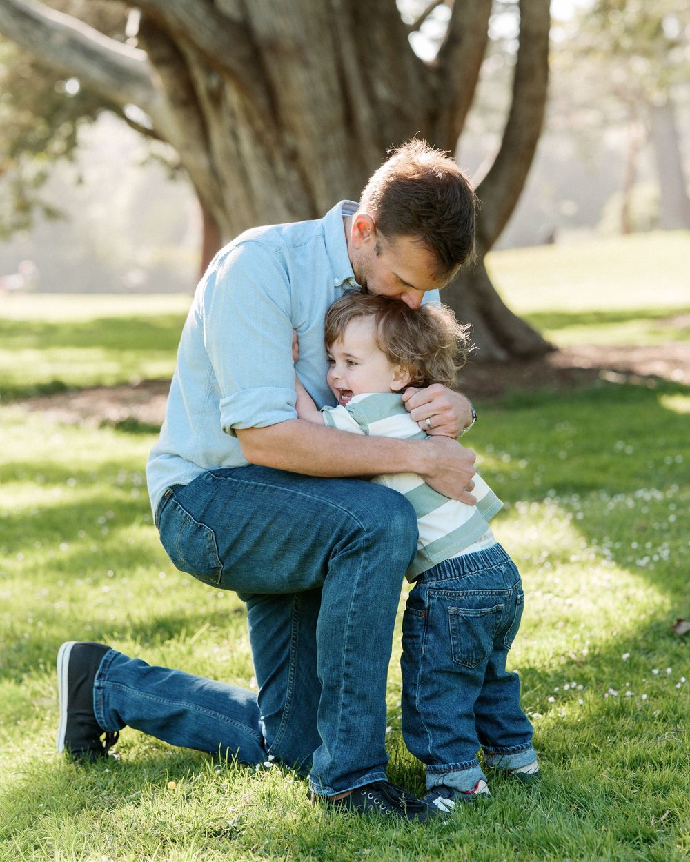 Father hugging his young toddler son on green grass in a sunlit park near a large tree.