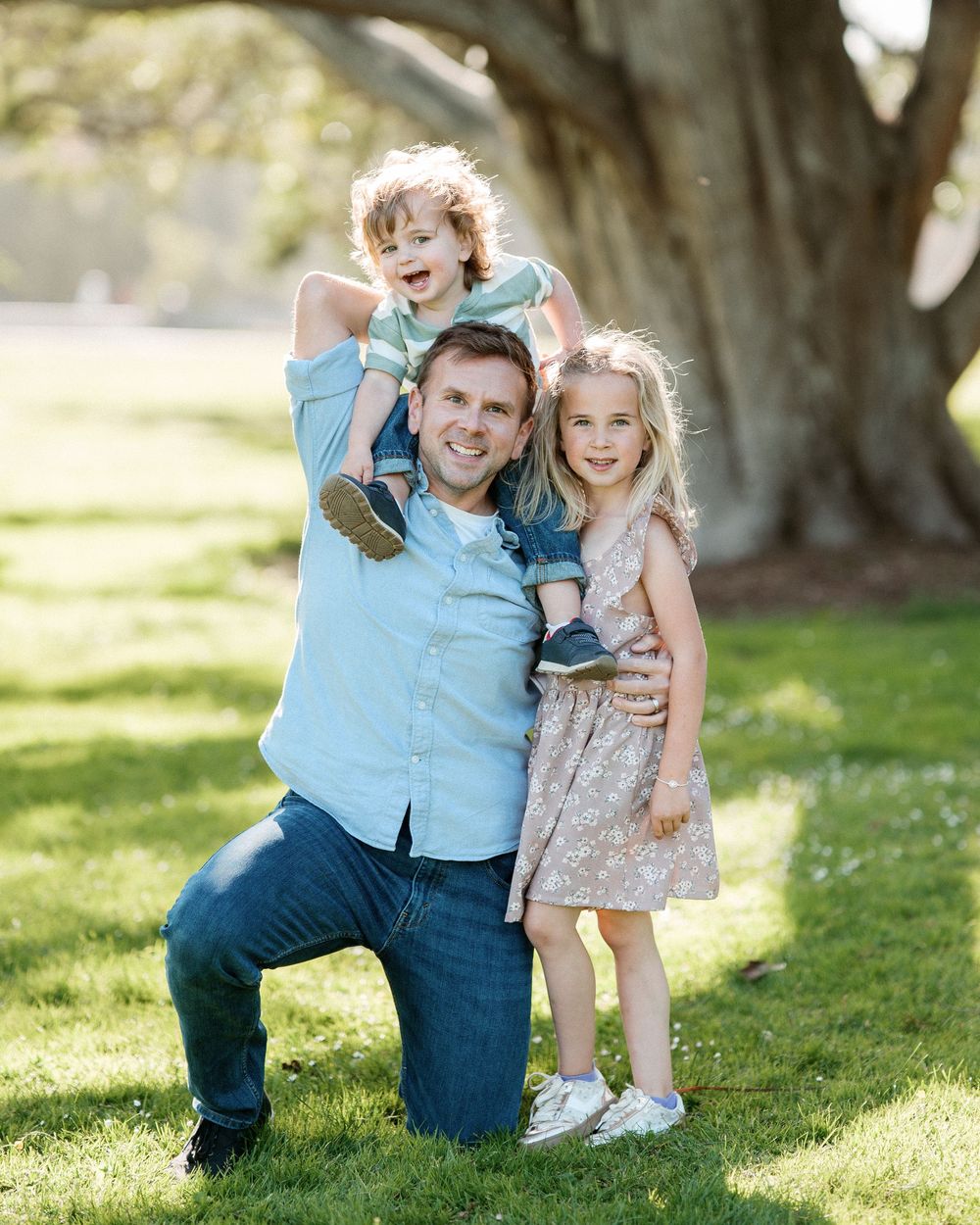 A smiling father kneels on green grass holding two young children near a large tree outdoors.