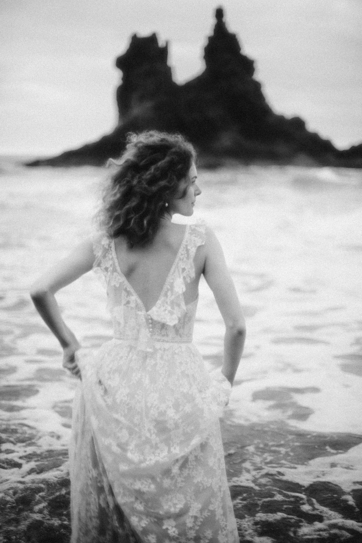 Black and white photo of a woman in a lace backless gown standing on rocky shore gazing at dramatic sea stacks.