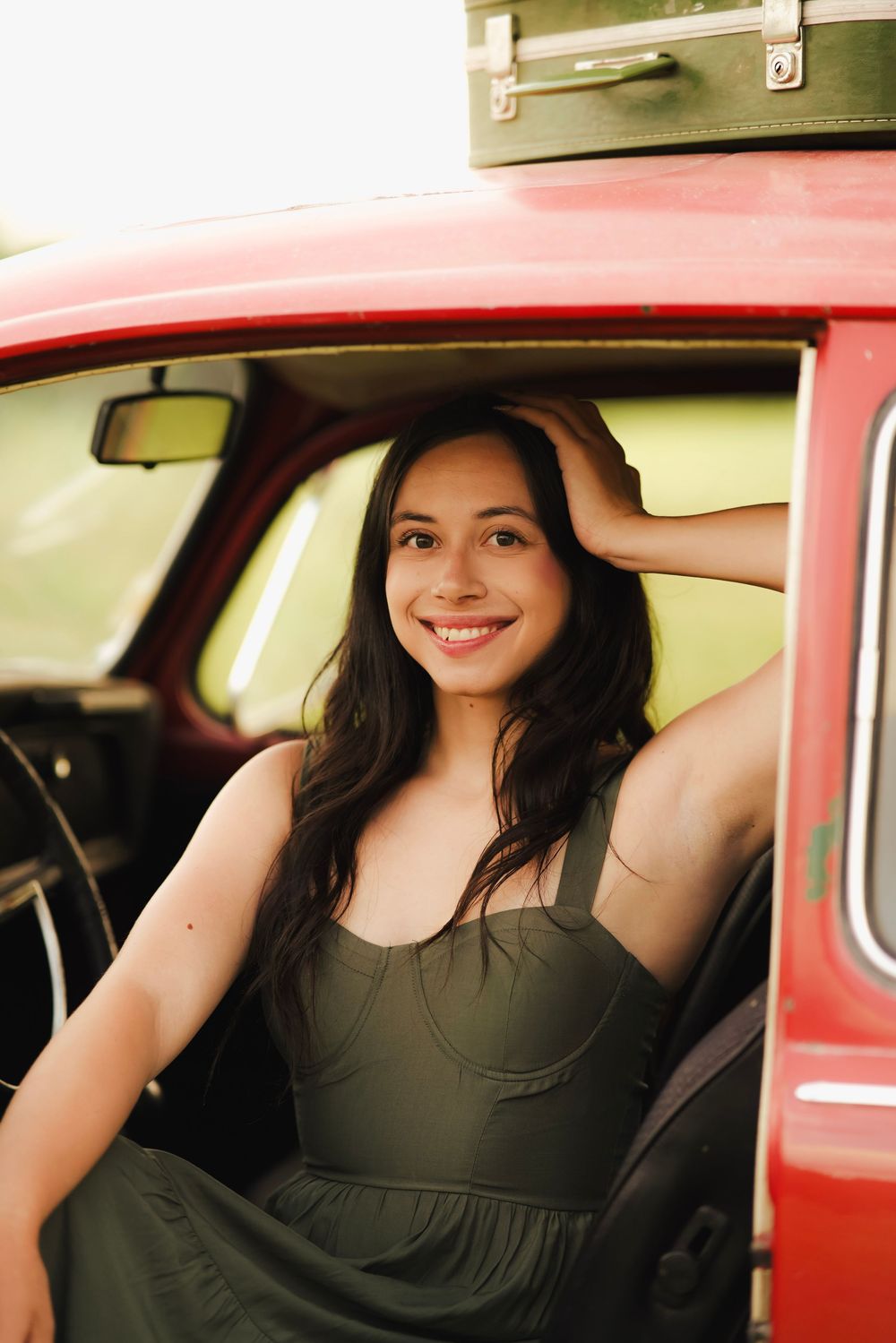 Smiling young woman with dark hair in olive dress sitting in vintage red car with suitcases on roof.