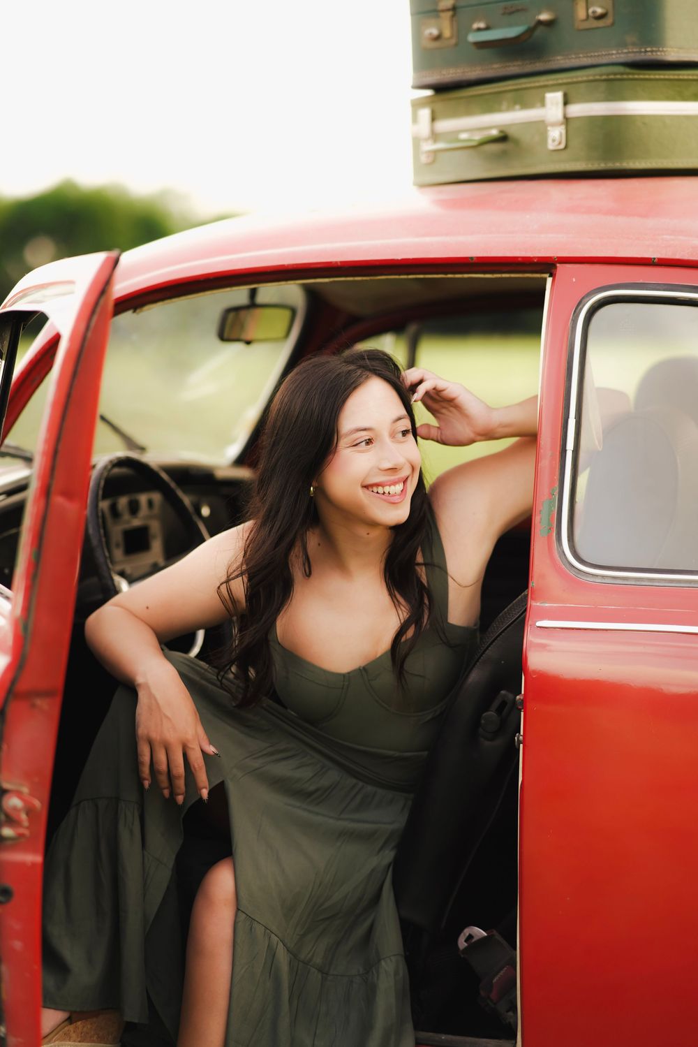 Happy young woman in olive green dress leaning out open door of classic red car with luggage on top.