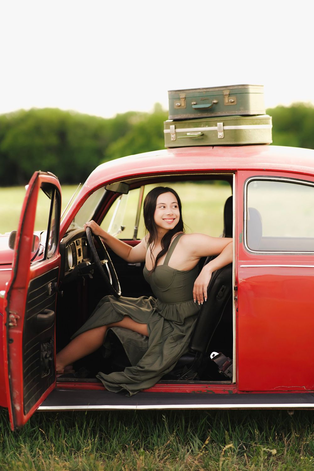 Smiling young woman in olive dress seated in open door of classic red car with vintage suitcases on roof.