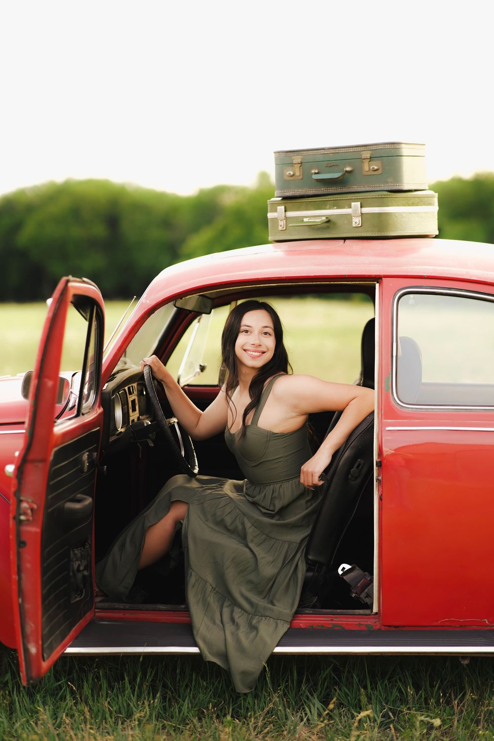 Smiling young woman in green dress sitting in open door of vintage red car with luggage stacked on roof.
