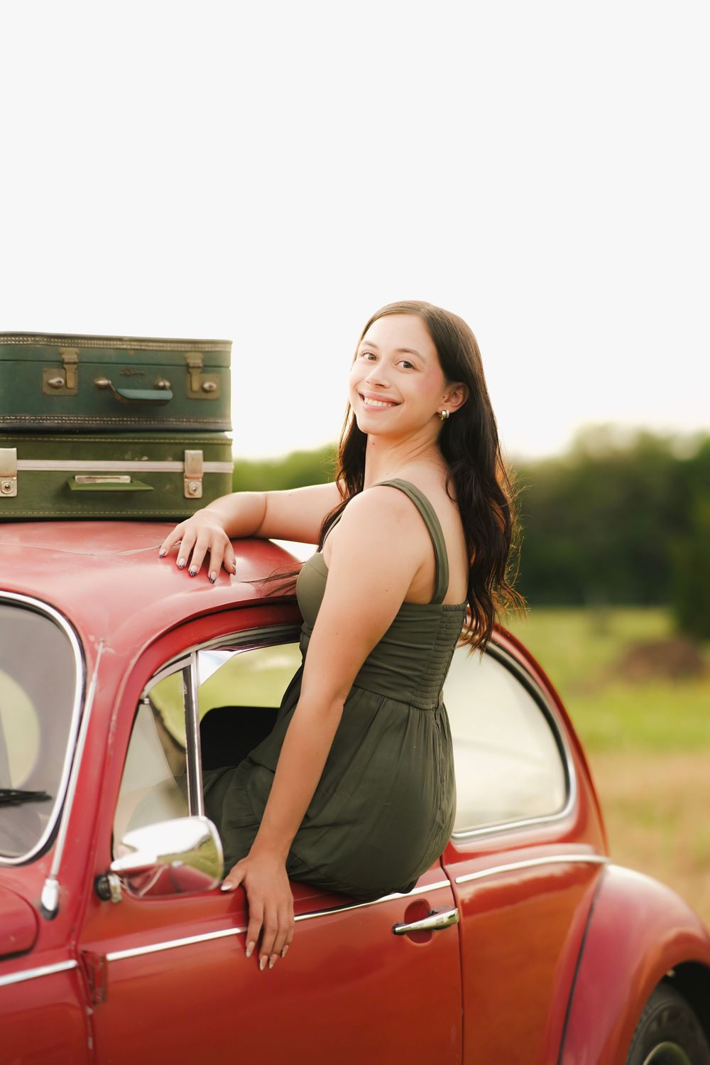 Smiling young brunette woman in olive dress leaning on classic red car with stacked vintage luggage on roof.