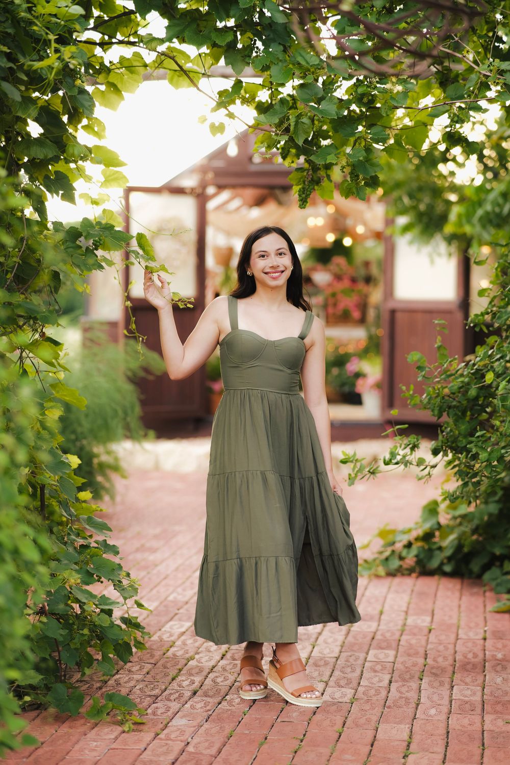 Smiling woman in olive green midi dress standing under lush green tree branches in a garden pathway.