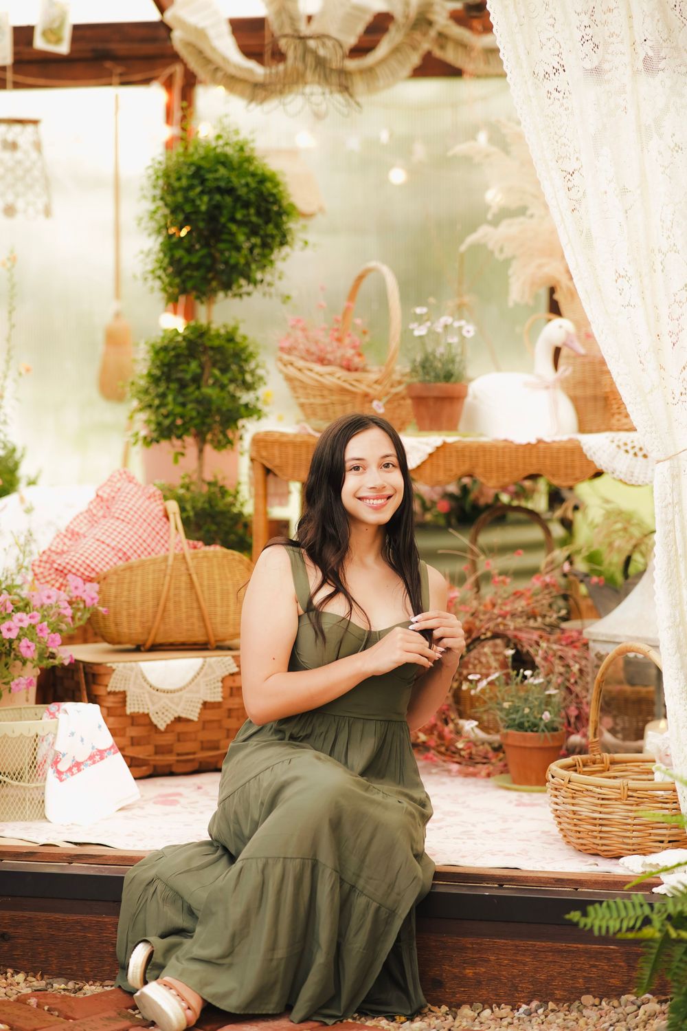 Young woman in olive dress sitting on porch surrounded by wicker baskets and potted plants smiling.