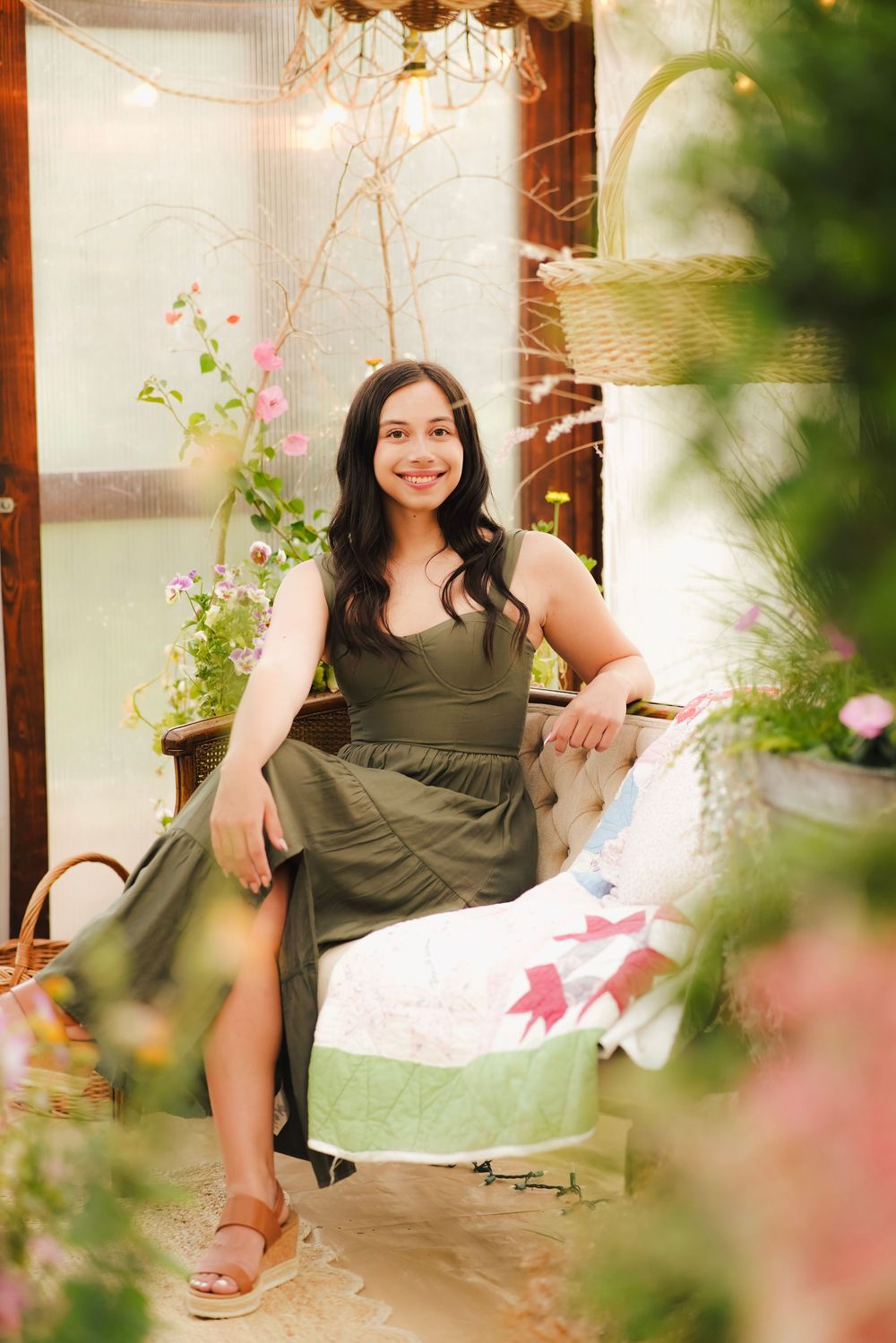Smiling woman in olive green dress seated on vintage bench surrounded by colorful flowers in greenhouse.