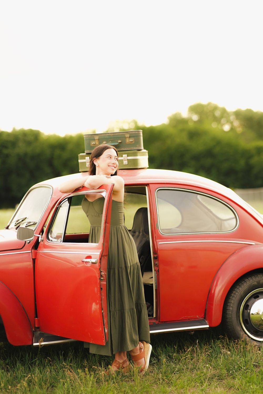 Smiling woman in olive dress leaning on red vintage VW Beetle with stacked suitcases on roof outdoors.