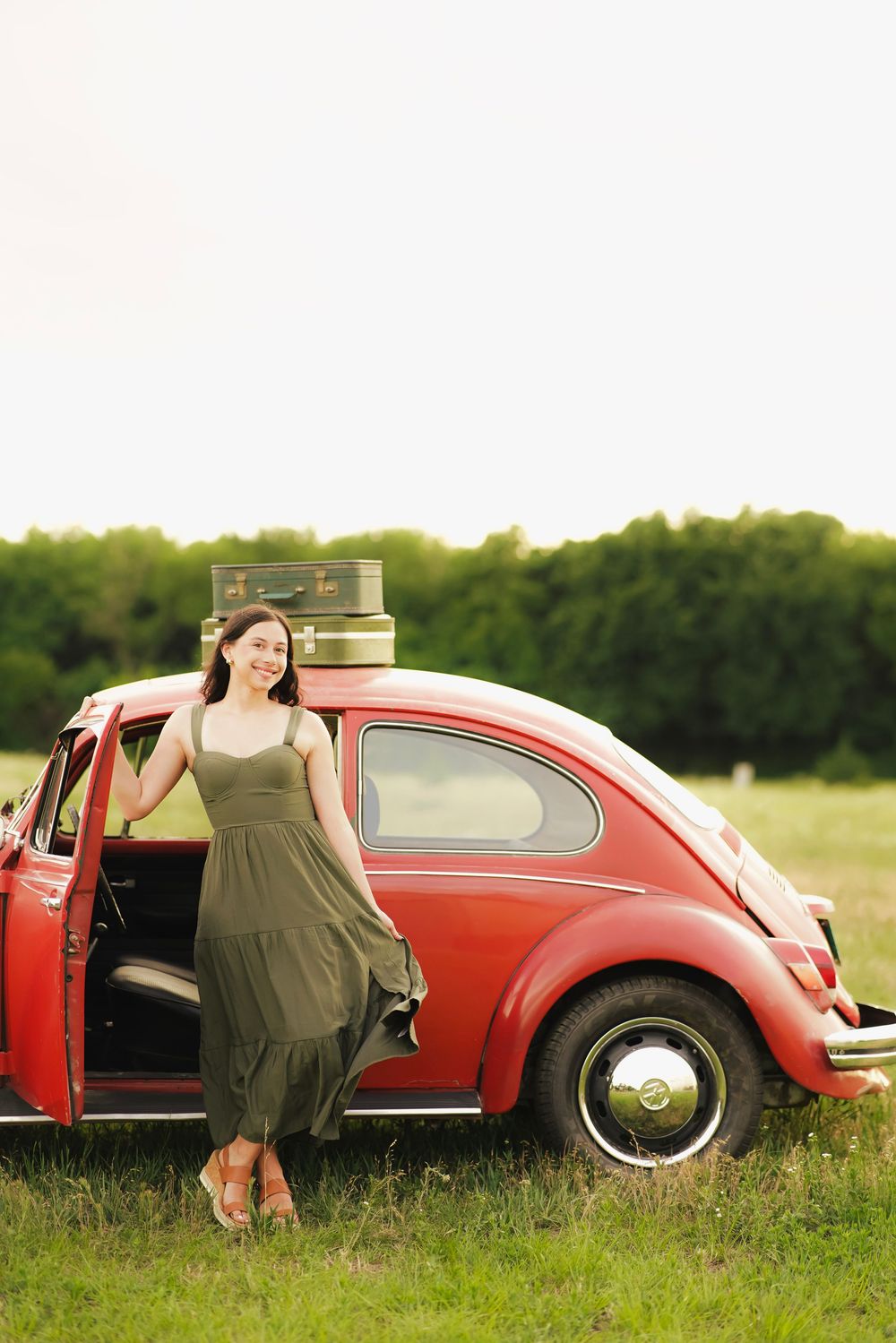 Woman in olive green midi dress standing beside a classic red VW Beetle with vintage luggage on top.
