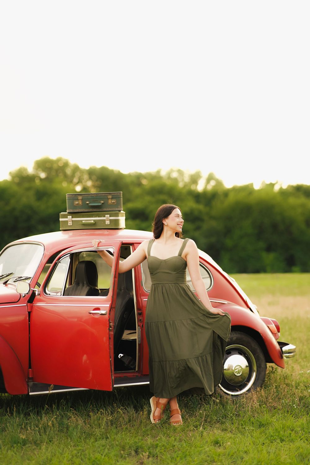 Woman in green dress posing beside a red vintage Volkswagen Beetle with suitcases on roof in field.