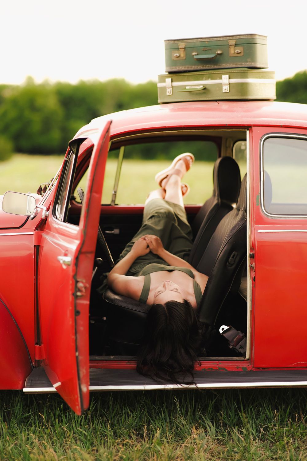 Young woman lying across vintage red car seat with feet up, luggage stacked on roof, door open in field.