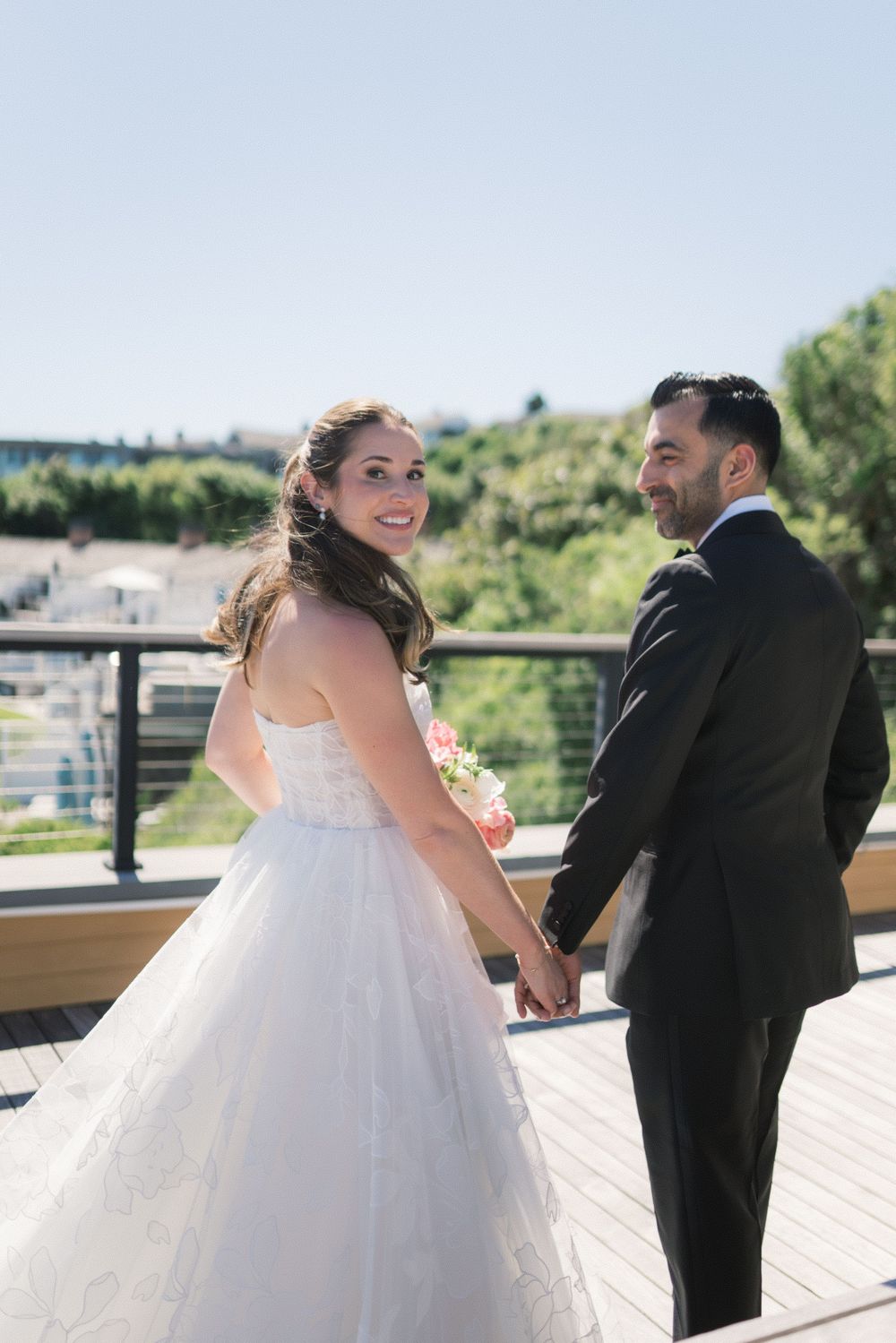 A smiling bride in a white ballgown holds hands with her groom on a sunny outdoor terrace.