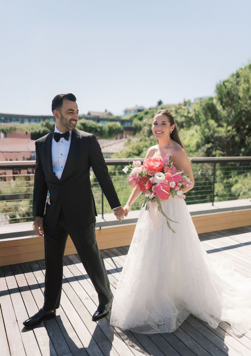 A happy bride and groom hold hands on a rooftop terrace, bride holding a vibrant pink bouquet.