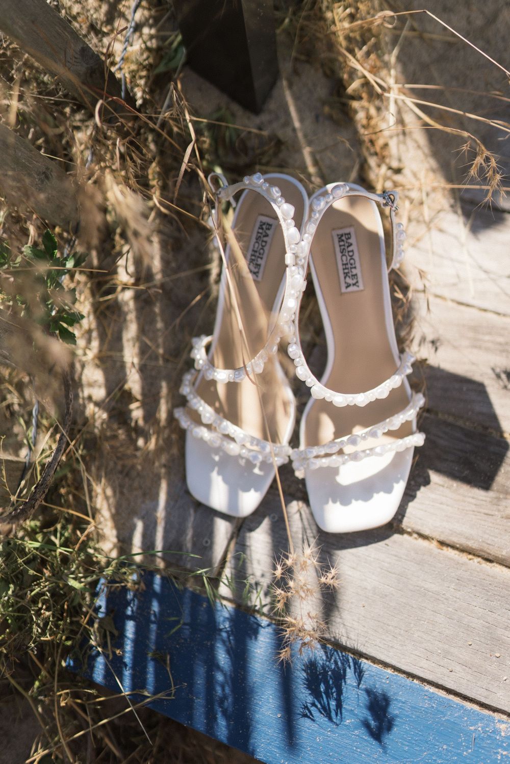 White pearl-embellished bridal heels by Badgley Mischka resting on wooden steps surrounded by natural dried grass.