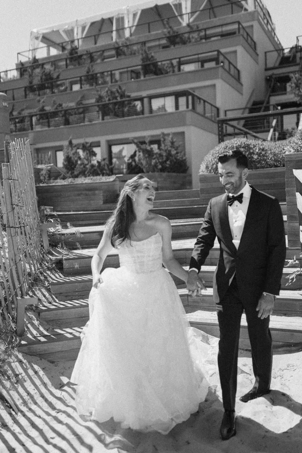 Black and white photo of bride and groom walking hand in hand on beach with modern hotel building backdrop.