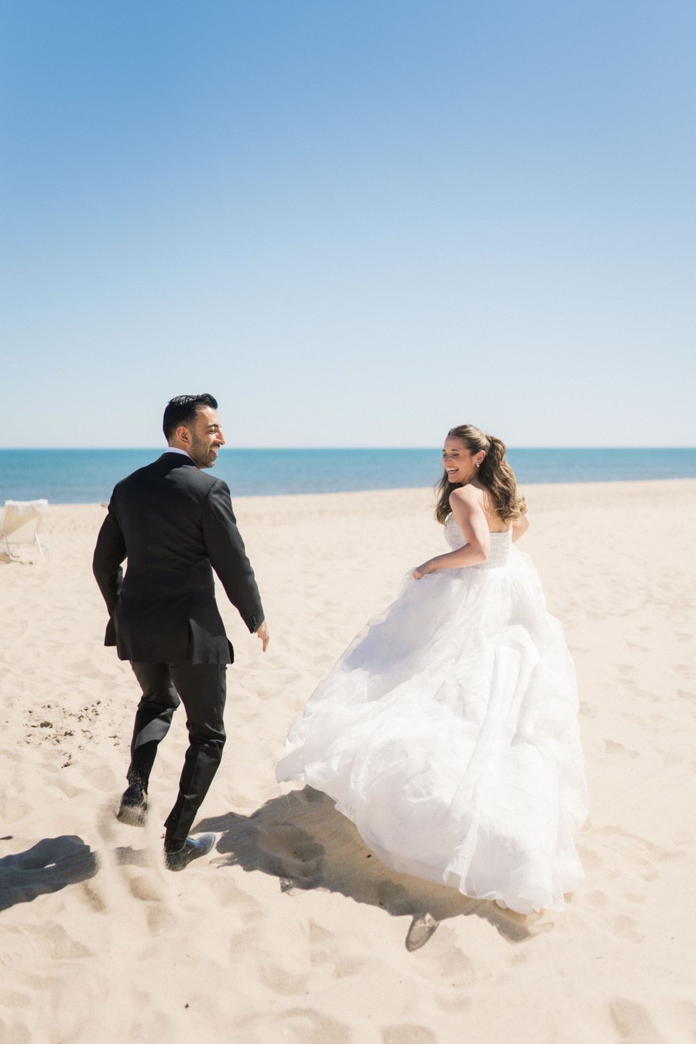 Joyful bride and groom running together on a sunny sandy beach with blue ocean in the background.