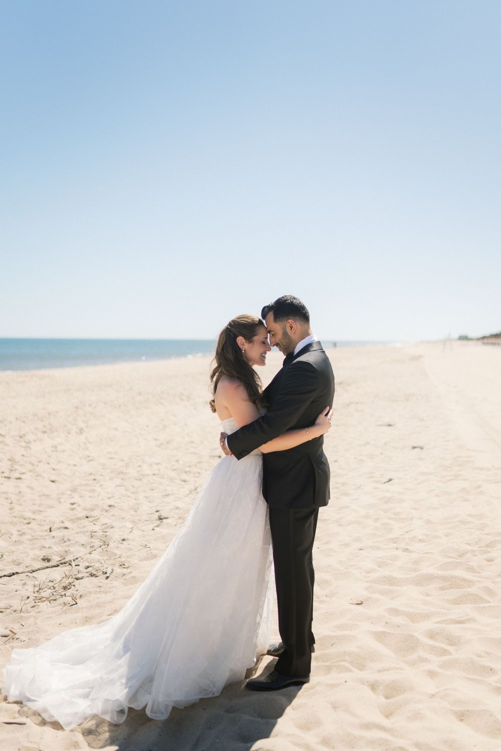 Bride and groom share romantic embrace on sandy beach with clear blue sky on wedding day.