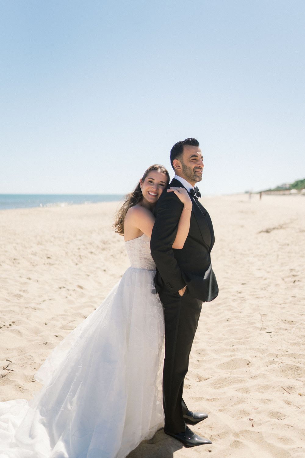 Happy bride embracing groom from behind on a sandy beach with clear blue sky and ocean view.