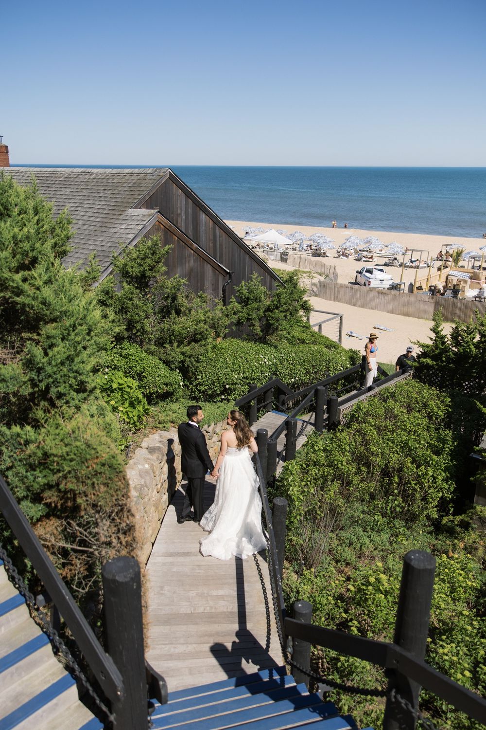 Bride and groom descend wooden staircase surrounded by lush greenery with ocean beach view behind them.