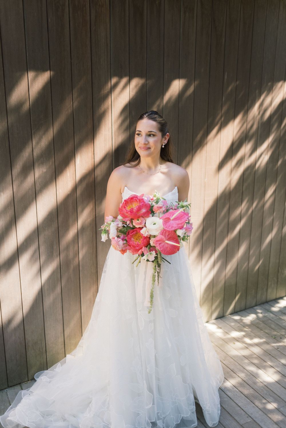 Bride in strapless white tulle gown holding vibrant pink and coral bouquet against wooden wall.
